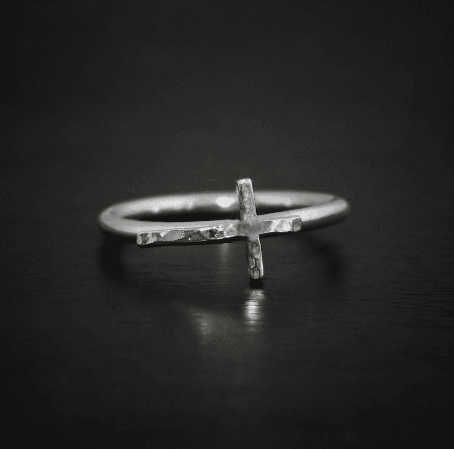 Close up of the "Creator" Ring. Made out of sterling silver. Ring has a thin, hammered oxidized cross going sideways across the ring band so that the top of the cross is on the right side of the ring band, the bottom of the cross is on the left side, and the two sides of the cross are sticking off of the ring band from the center of the cross. Ring is sitting against a dark background.